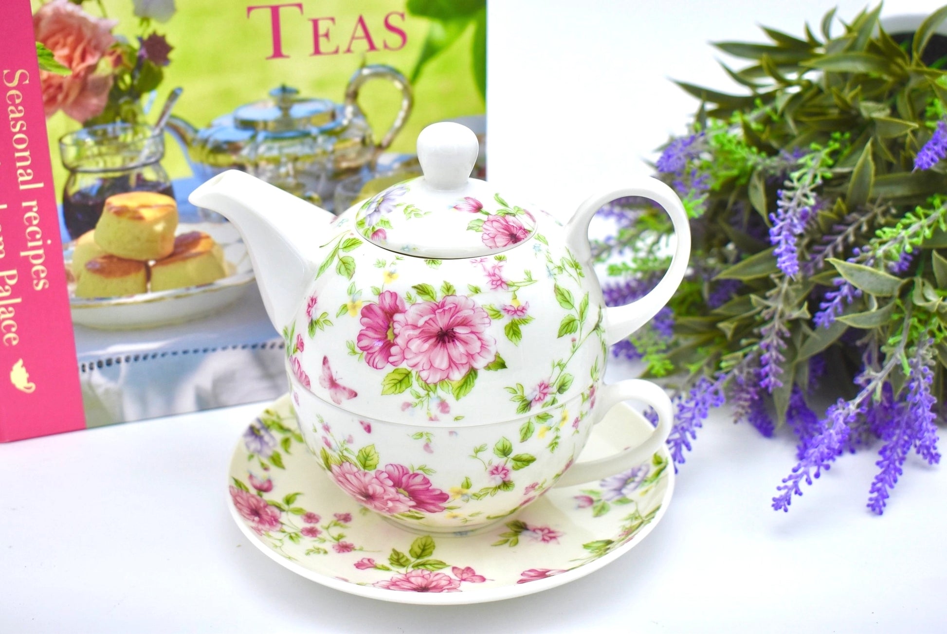 Decorative teapot with floral pattern on a saucer, next to a book titled 'Seasonal Recipes' and purple flowers.